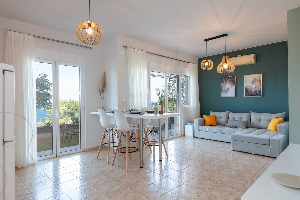 A modern living room with a dining area, a grey sofa with orange cushions, and a large window with curtains.
