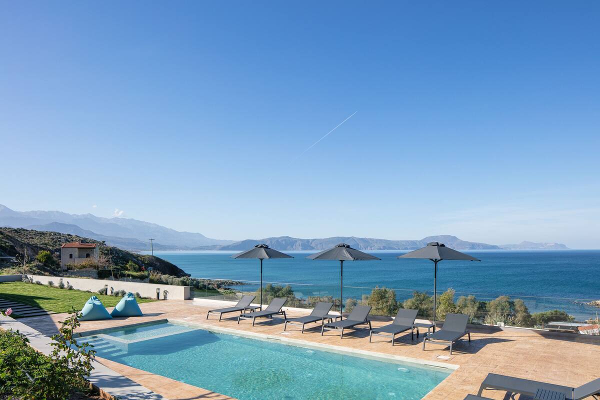 A poolside area with lounge chairs and umbrellas, overlooking a body of water and mountains.