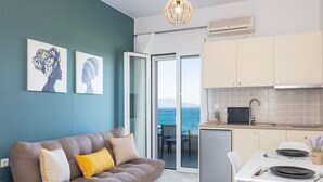 A modern living room with a sofa, a dining table set for two, and a kitchen area with a view of the sea. - Pietra Di Mare (Rethymno)