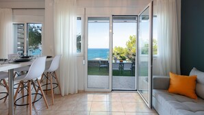 A modern living room with a sofa, a dining table with chairs, and a view of the sea through large sliding glass doors. - Pietra Di Mare (Rethymno)