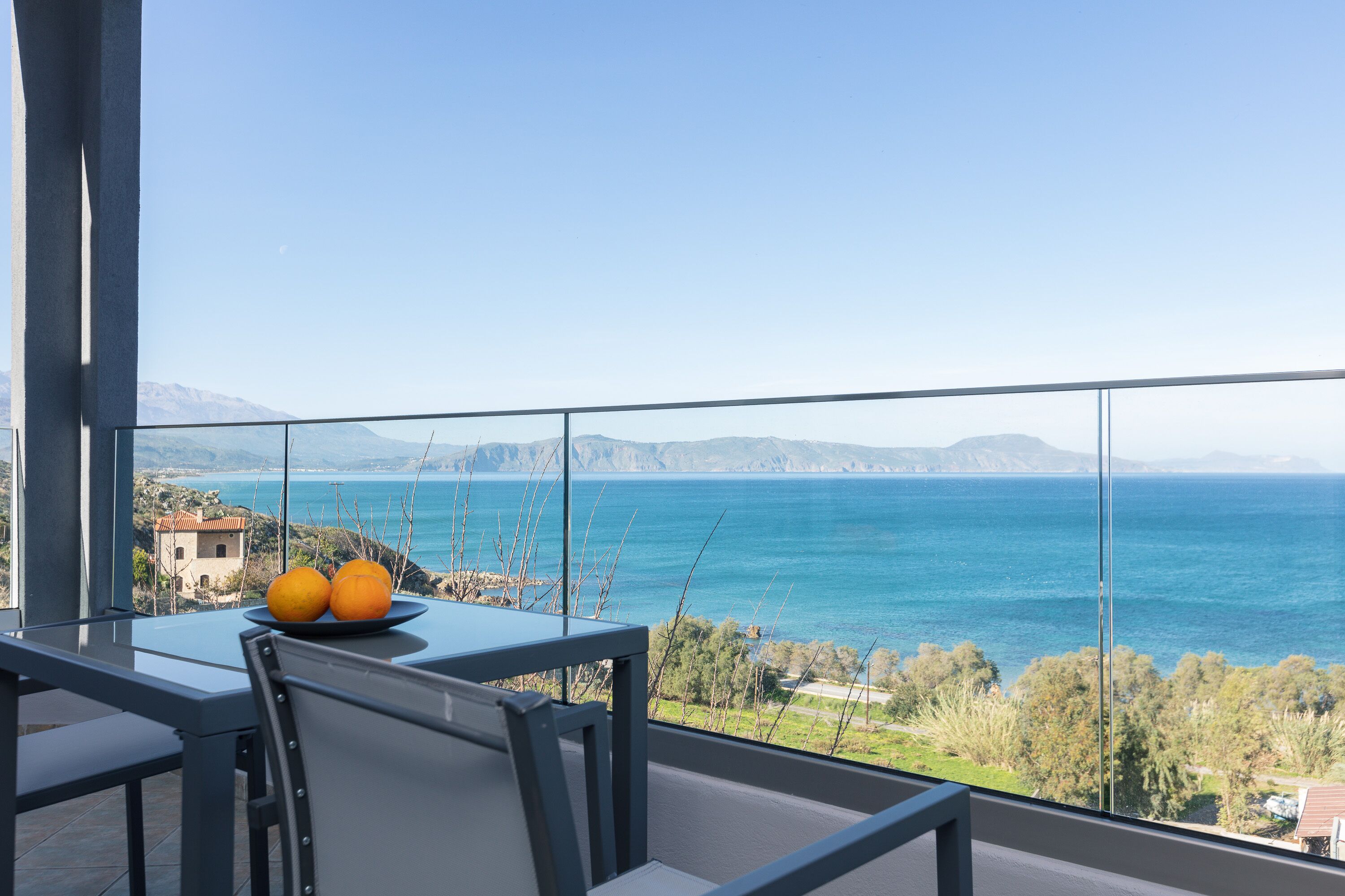 Een balkon met een tafel en stoelen, met uitzicht op de kust, bergen en een waterlichaam.