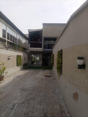 A courtyard with a paved walkway, a building with a glass entrance, and potted plants along the walls.
