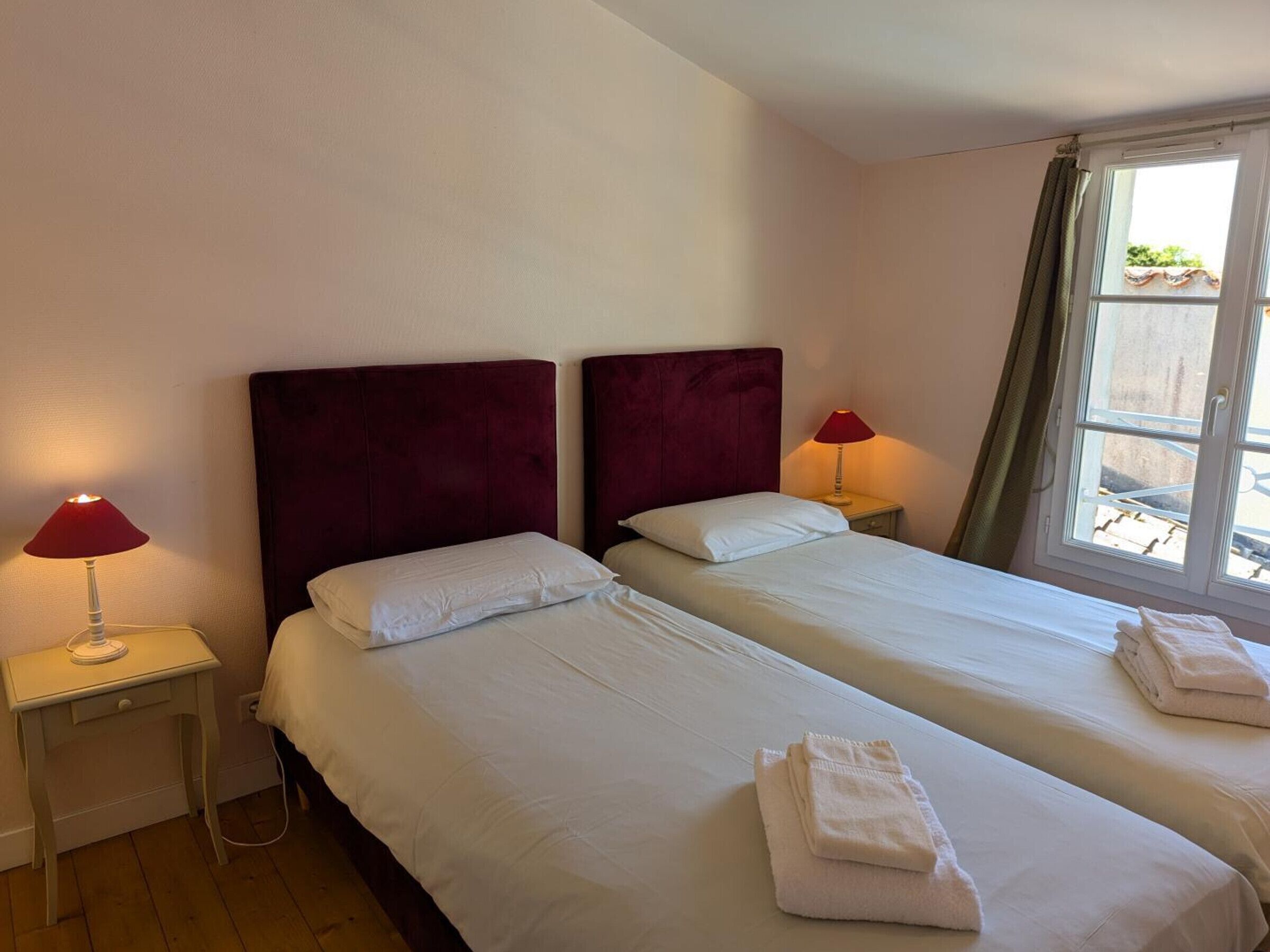 A hotel room with two beds, each with a red headboard, white bedding, and a bedside lamp. There is a window with a view of the outside.