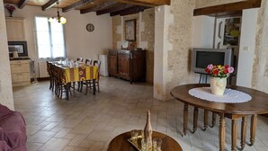 Un intérieur rustique avec des poutres apparentes, un coin repas avec une nappe jaune, une table ronde en bois sur laquelle se trouve un plateau, et un téléviseur en arrière-plan.