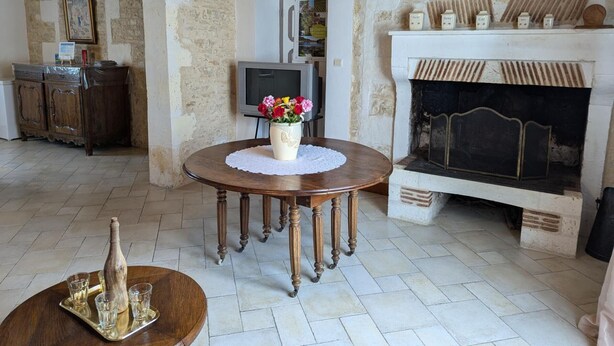 Un salon chaleureux avec une cheminée, une table ronde en bois ornée d’un vase de fleurs et un téléviseur en arrière-plan.