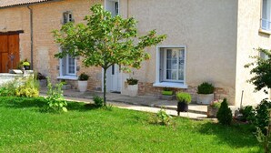Un bâtiment à deux étages avec une façade en pierre, un petit arbre dans le jardin devant et des plantes en pot sur les marches.