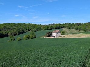 Un paysage rural avec un bâtiment en pierre, entouré de champs verdoyants et d’arbres, sous un ciel bleu dégagé.