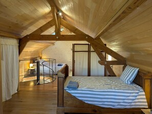An attic bedroom with a sloped ceiling, a bed with striped bedding, a small desk with a lamp, and a window.