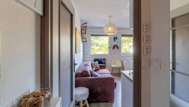 A narrow hallway leading to a room with a bed, a small stool, and a window with a view of greenery. - Appartement au Pied des Pistes (Arâches-la-Frasse)