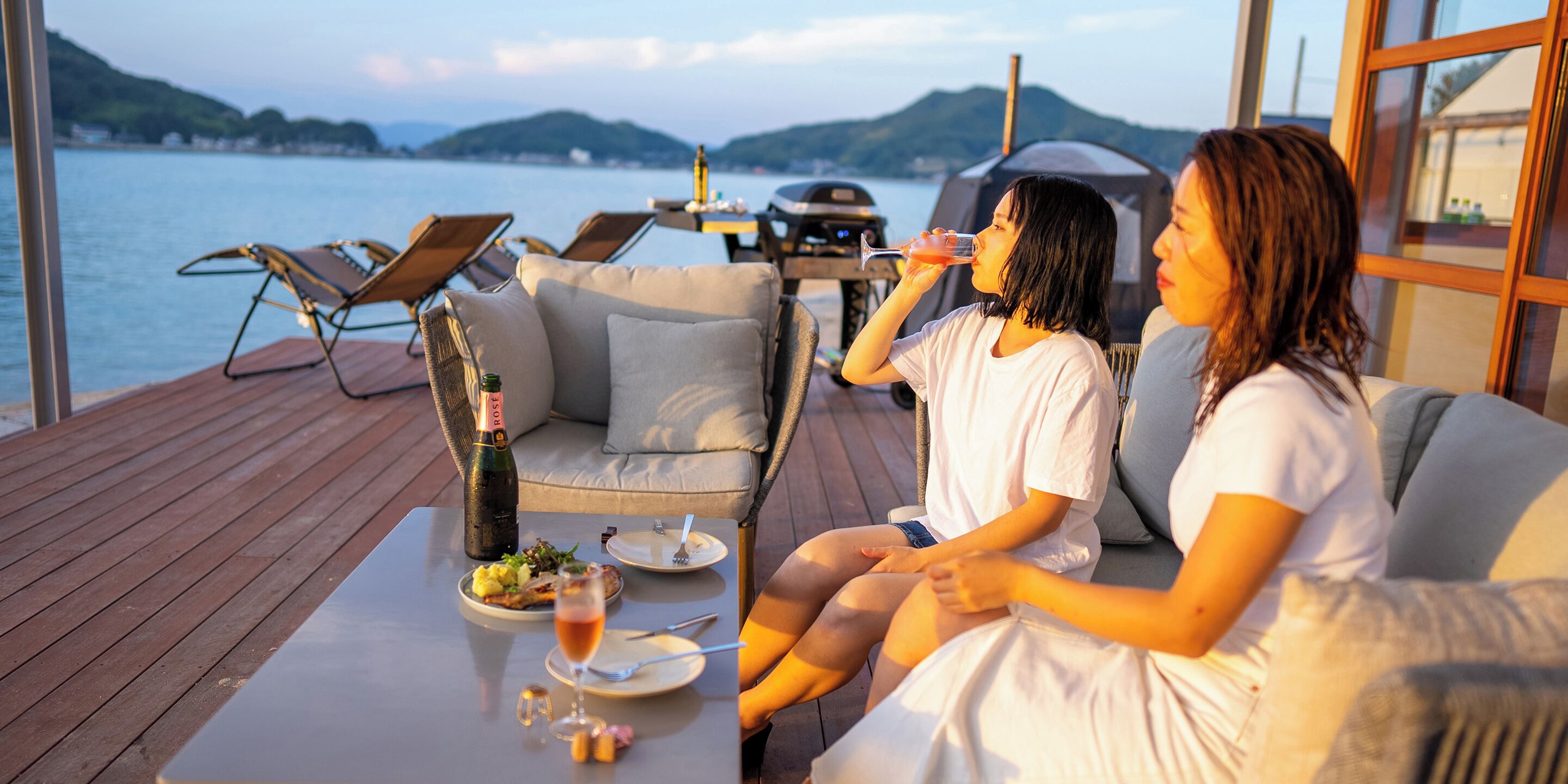 A couple relaxing on a deck with a table set for a meal, overlooking a body of water.