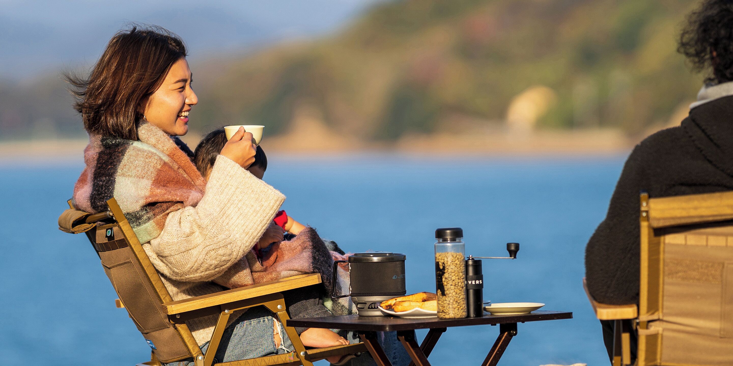 A person seated at a table by a body of water, with a portable stove and food items on the table.