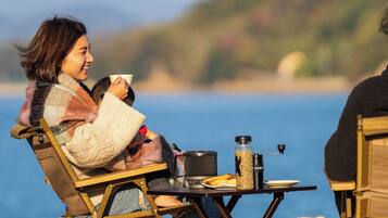 A person seated at a table by a body of water, with a portable stove and food items on the table.