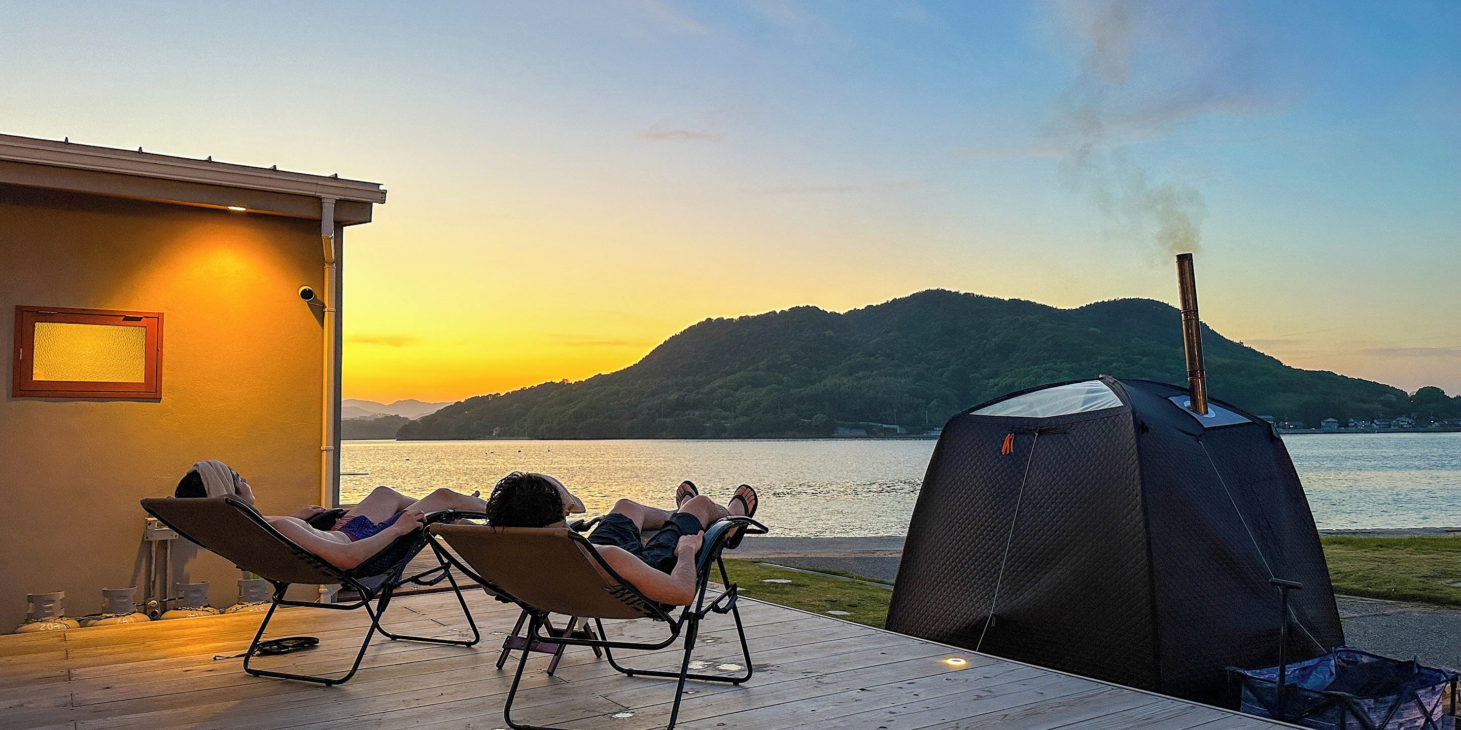 A deck with lounge chairs overlooking a lake, a tent, and a building with a window.