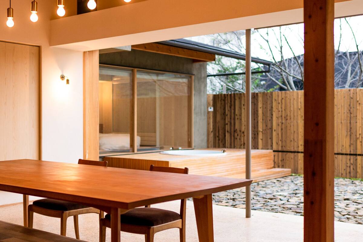 A modern dining area with a wooden table and chairs, large windows, and a view of a wooden deck and outdoor space.
