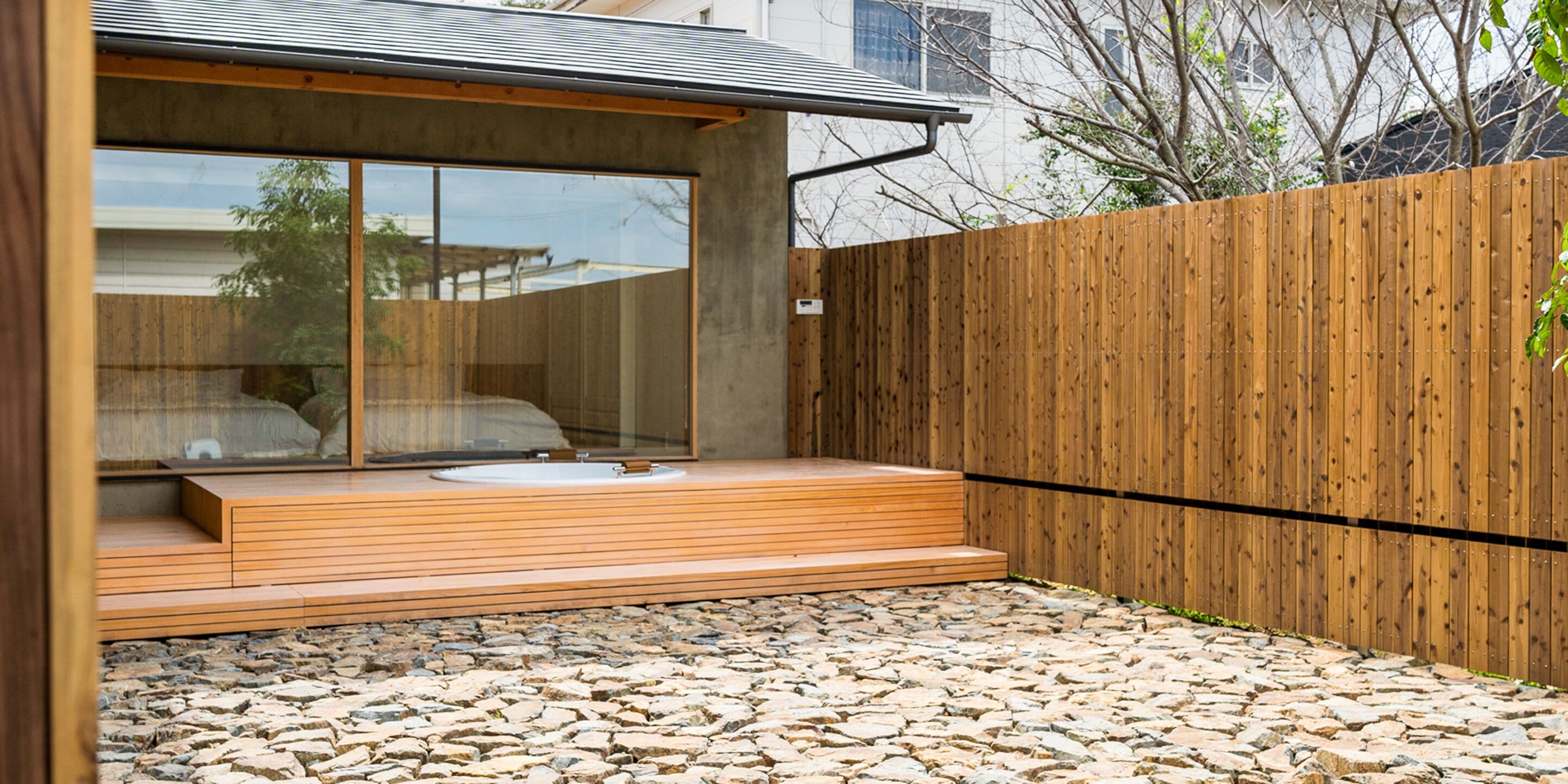 A modern outdoor bathroom with a stone-tiled floor, wooden deck, and a large window.