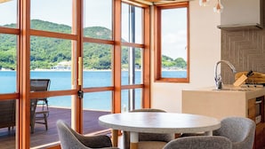 A room with a large window overlooking a body of water, a round table, and a kitchen area with a sink and stove. - Setouchi Hideaway Resort (Onomichi)