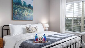 Een netjes opgemaakt bed met witte lakens, een decoratief kussen en een dienblad met twee flessen Fiji water.