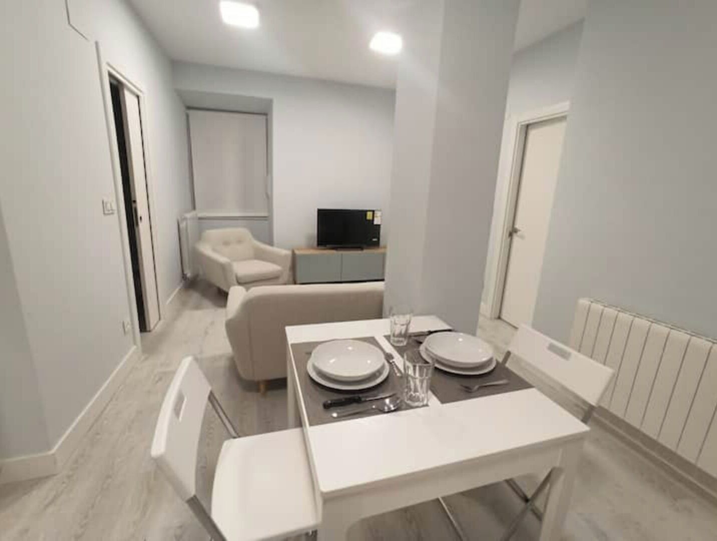A modern, compact living space with a dining area, a sofa, and a television in the background.