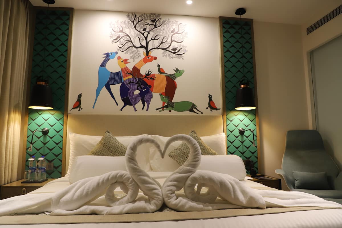 Habitación de hotel con cama, una toalla dispuesta en forma de corazón, un mural de animales en la pared y dos lámparas de noche.