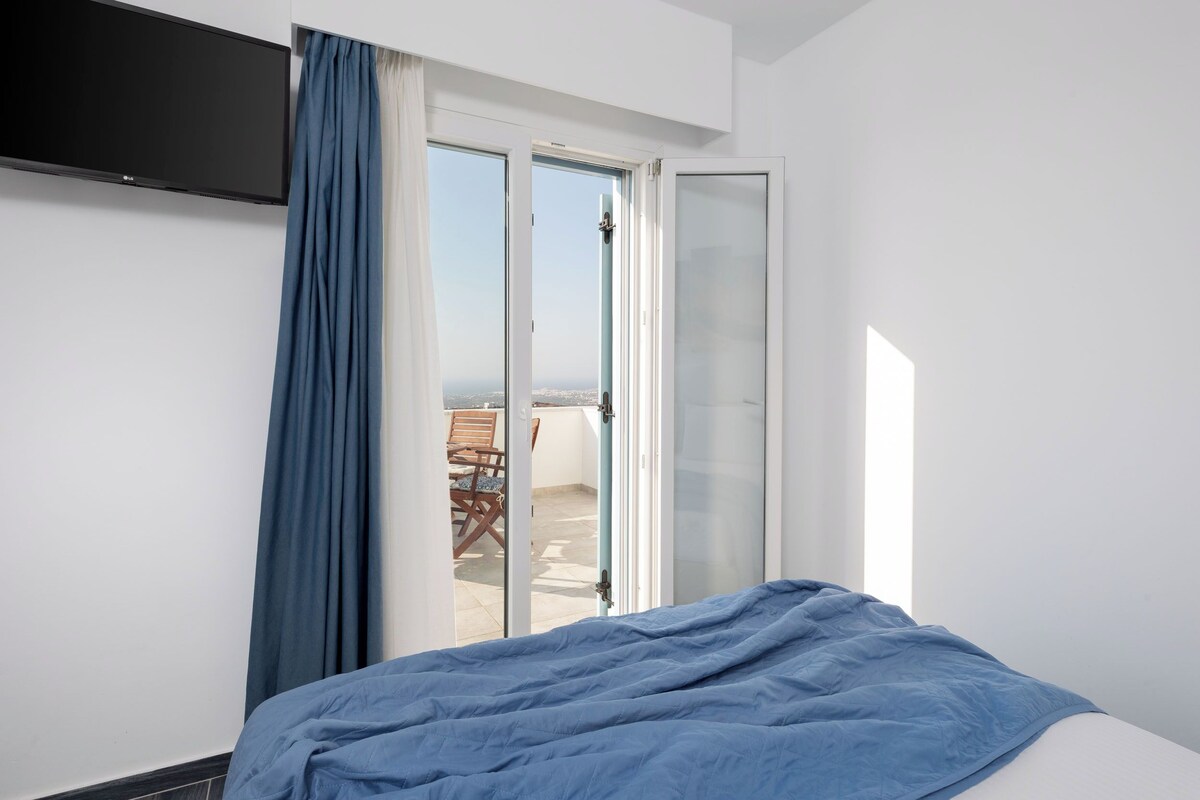 A bedroom with a bed, a TV mounted on the wall, and a sliding glass door leading to an outdoor seating area.