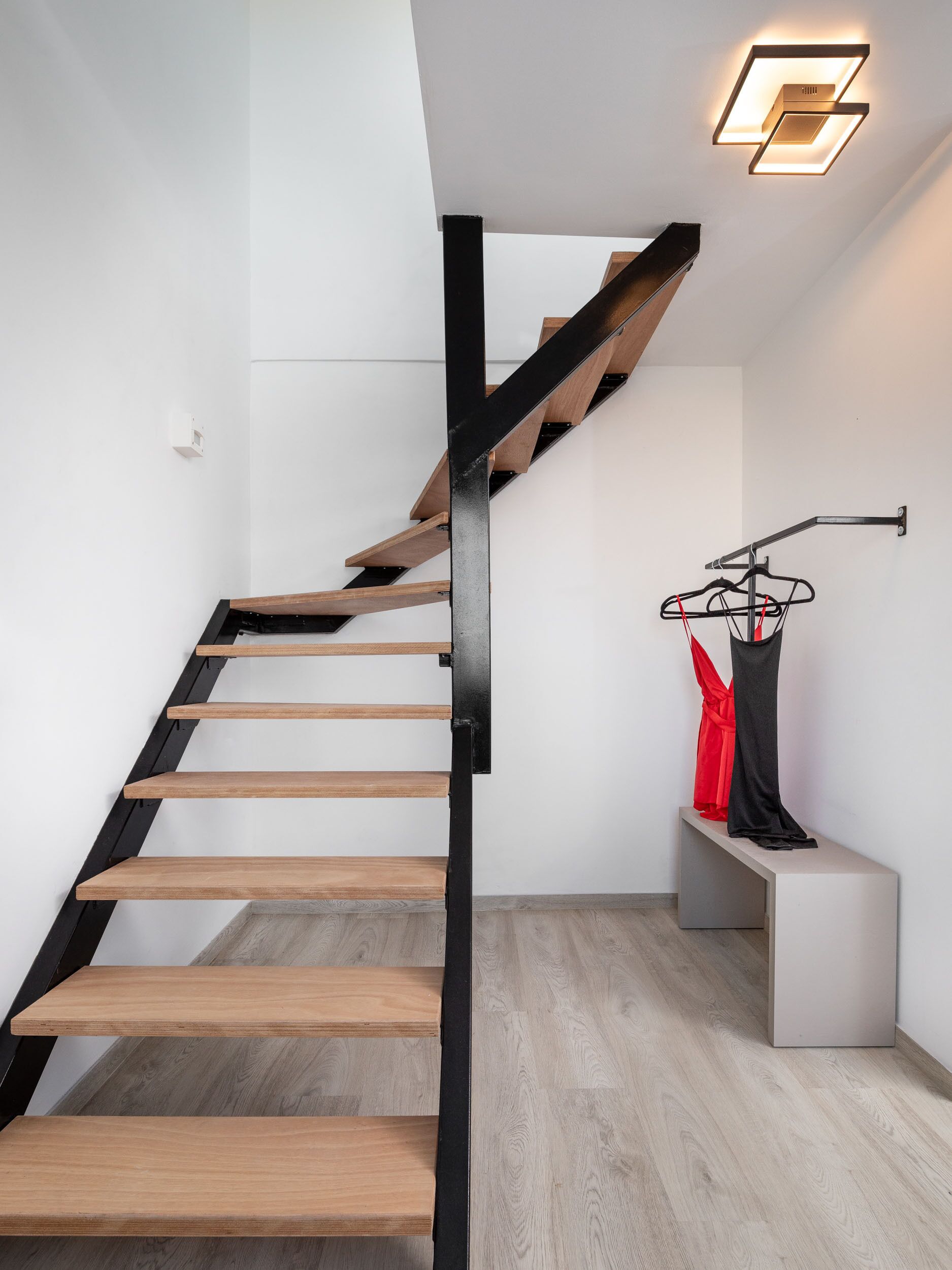 A modern staircase with wooden steps and black metal railings, leading to a landing with a wall-mounted light fixture and a bench with red items on it.