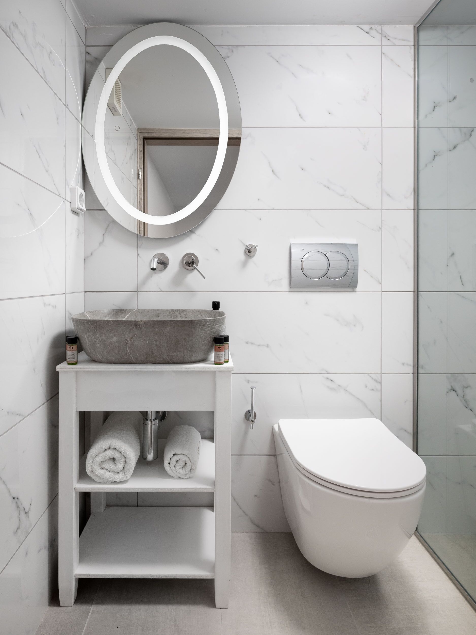 A modern bathroom with a marble-tiled wall, a round mirror, a vessel sink, a toilet, and a towel shelf.