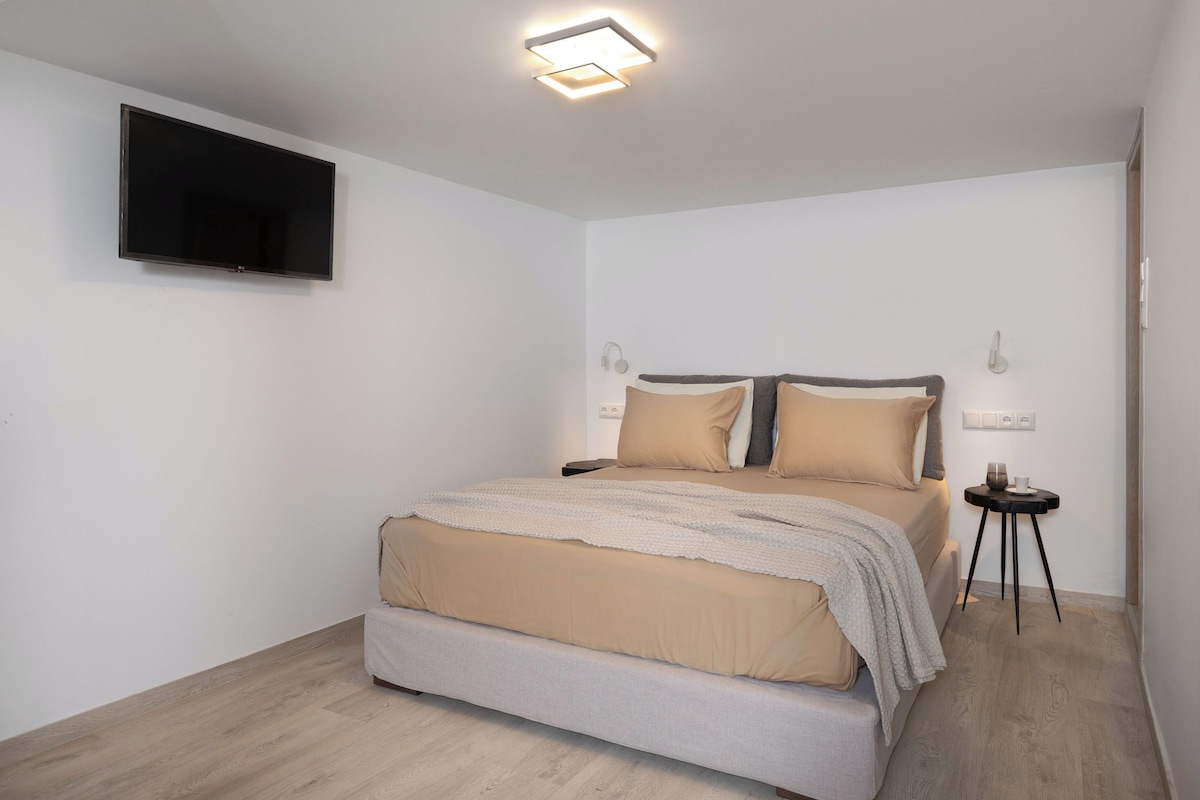 A modern bedroom with a large bed, a flat-screen TV mounted on the wall, and a small bedside table with a lamp.