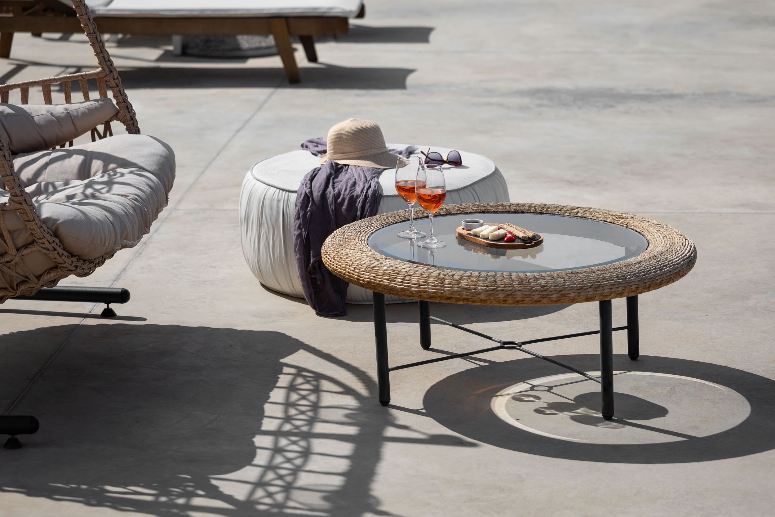 A round wicker table with a glass of drink and a plate of food, a cushioned chair, and a small round table with a hat and sunglasses.