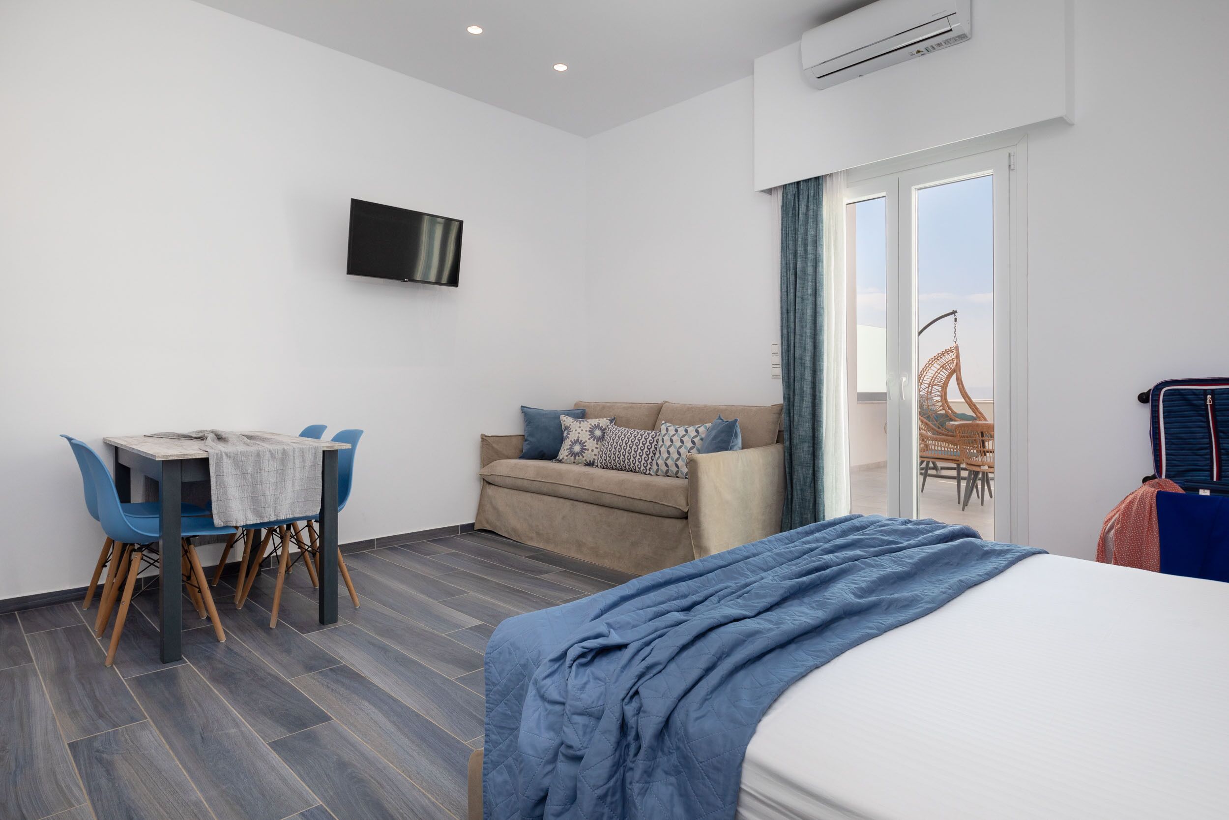 A modern hotel room with a bed, a sofa, a dining table with chairs, a TV mounted on the wall, and a balcony with a chair.