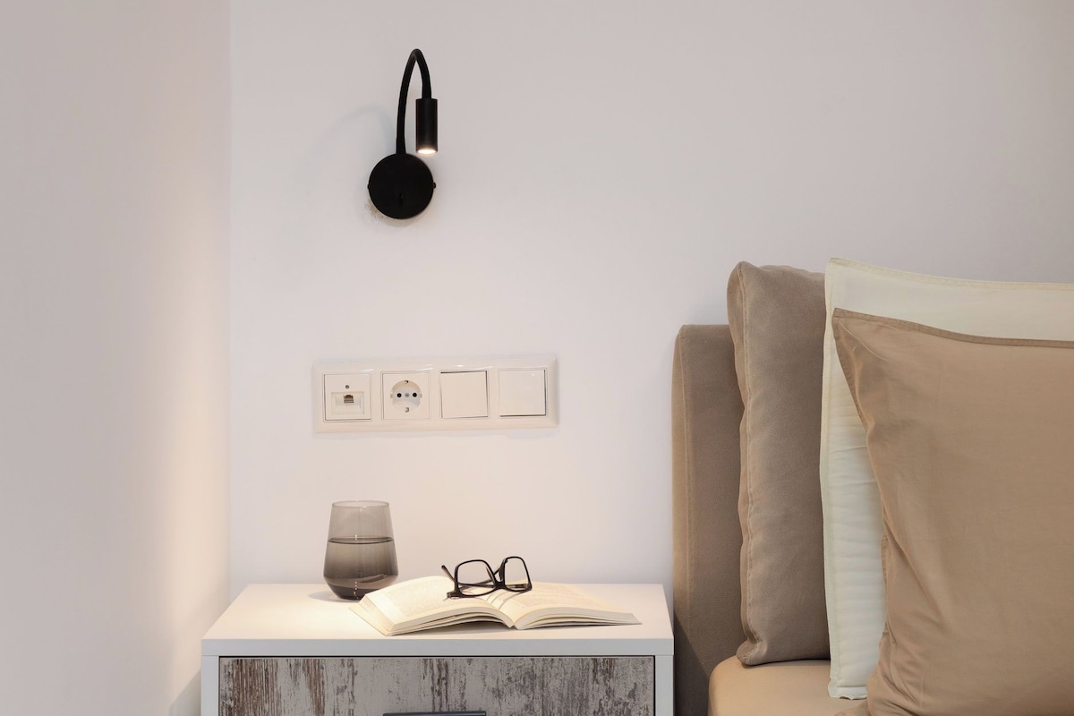 A modern bedroom with a white nightstand, a wall-mounted lamp, and a bed with beige bedding.