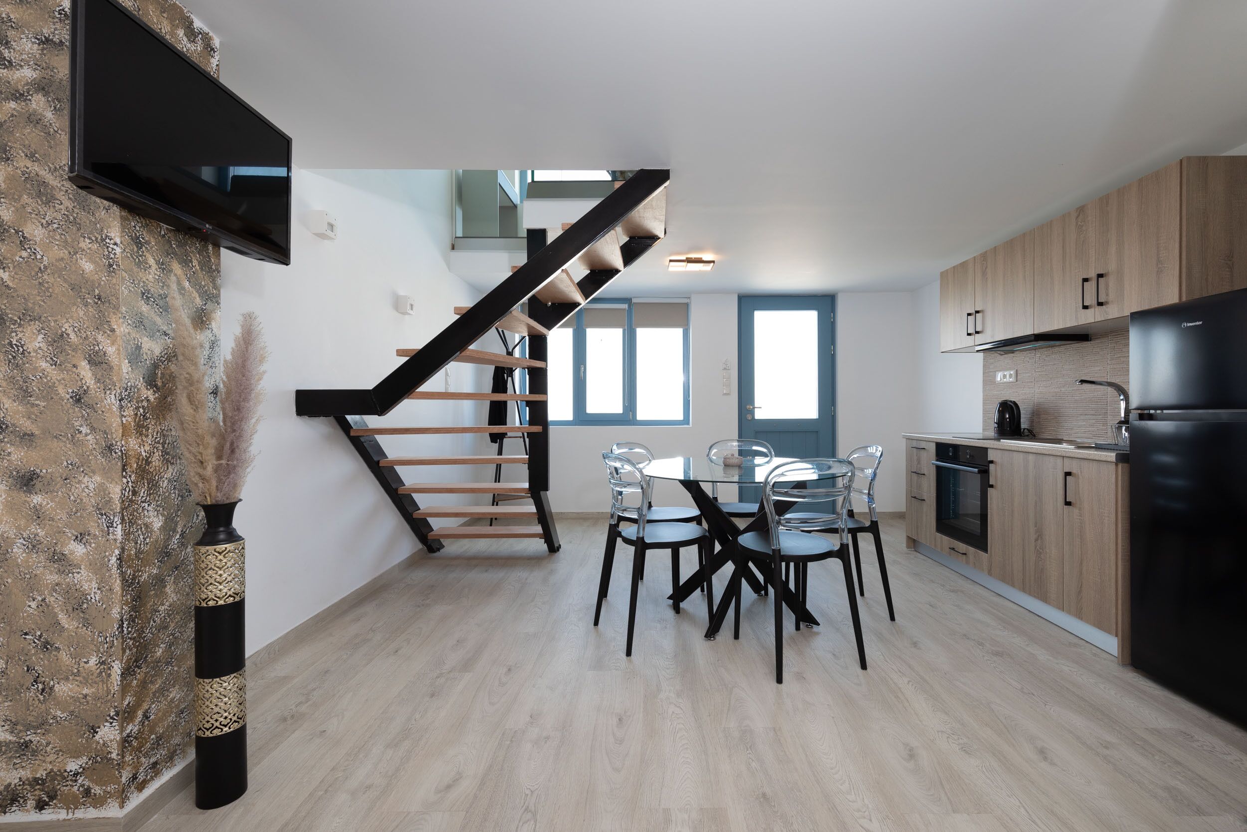 Une cuisine moderne avec des meubles en bois, un réfrigérateur noir et un coin repas comprenant une table et des chaises en verre.