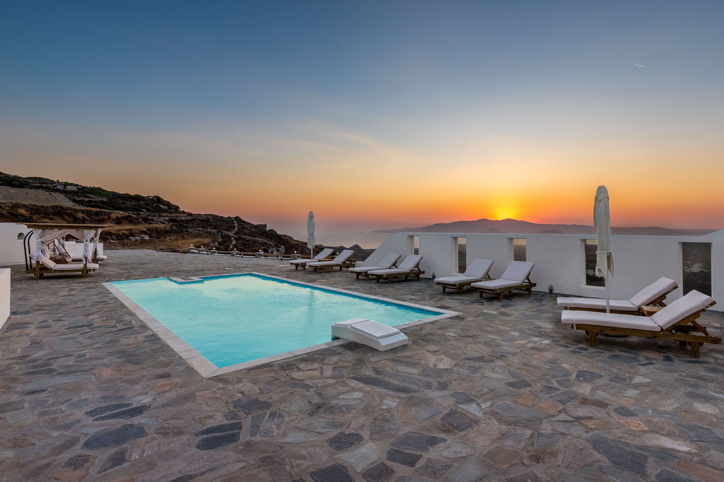 Un espace piscine doté d’un sol en pierre, de chaises longues blanches et offrant une vue sur le coucher de soleil.