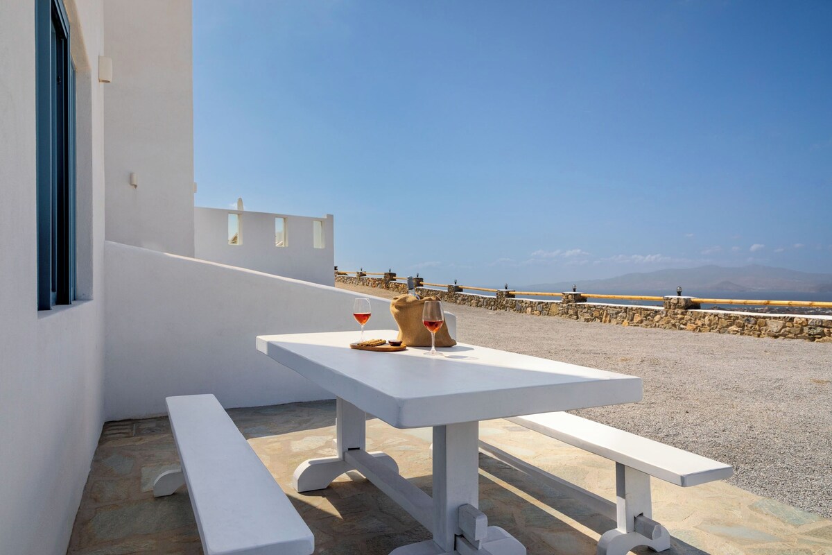 A white table with two wine glasses and a bag on a terrace with a stone floor and a stone wall.