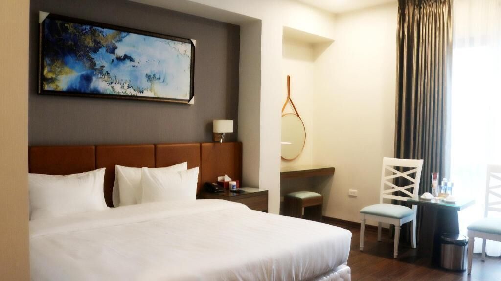 A hotel room with a large bed, a desk, a chair, a small table, a mirror, and a painting on the wall.