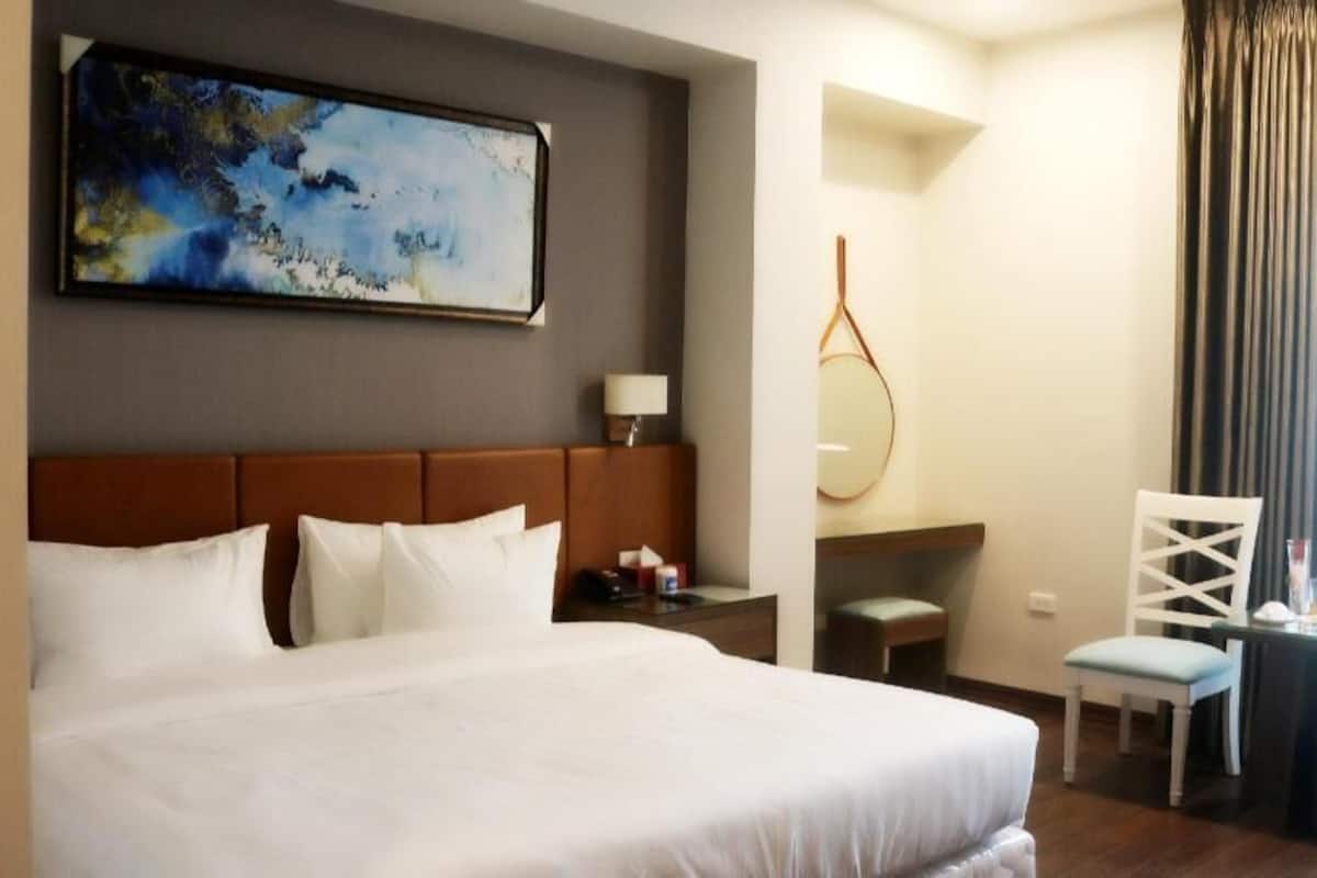 A hotel room with a large bed, a desk, a chair, a small table, a mirror, and a painting on the wall.