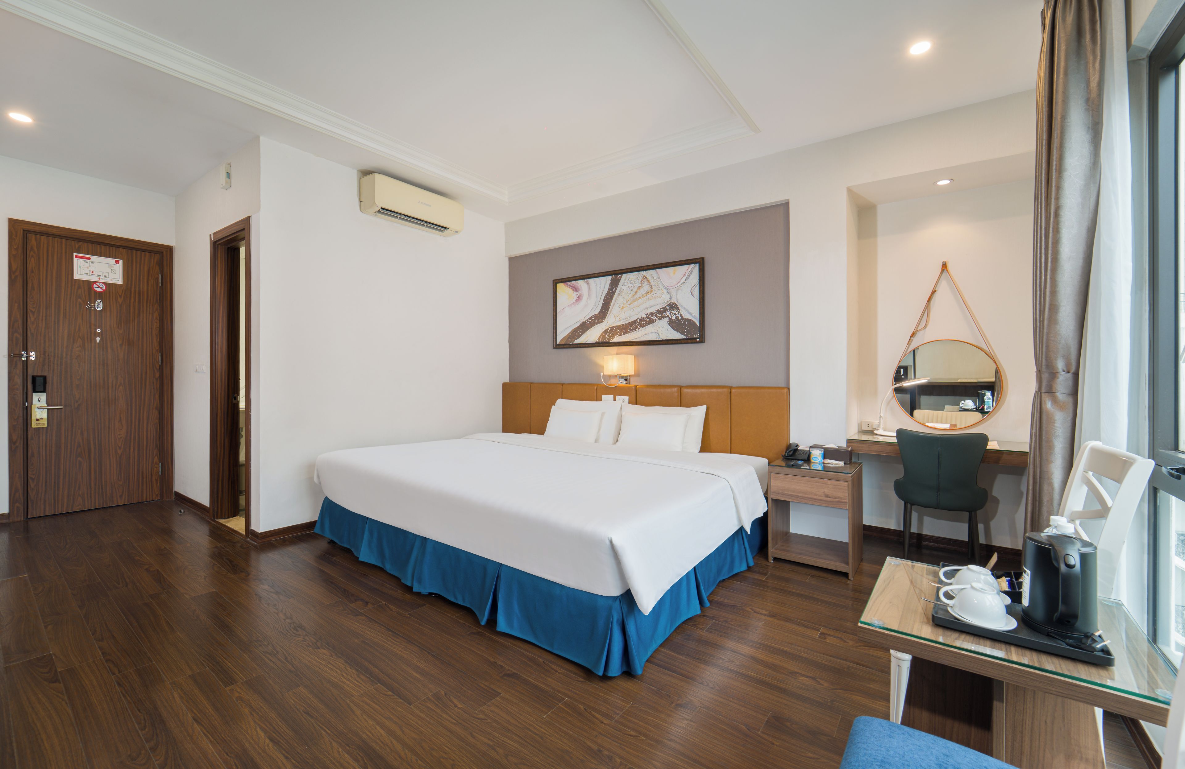 A hotel room with a large bed, a desk with a chair, a desk lamp, a coffee machine, and a window with curtains.