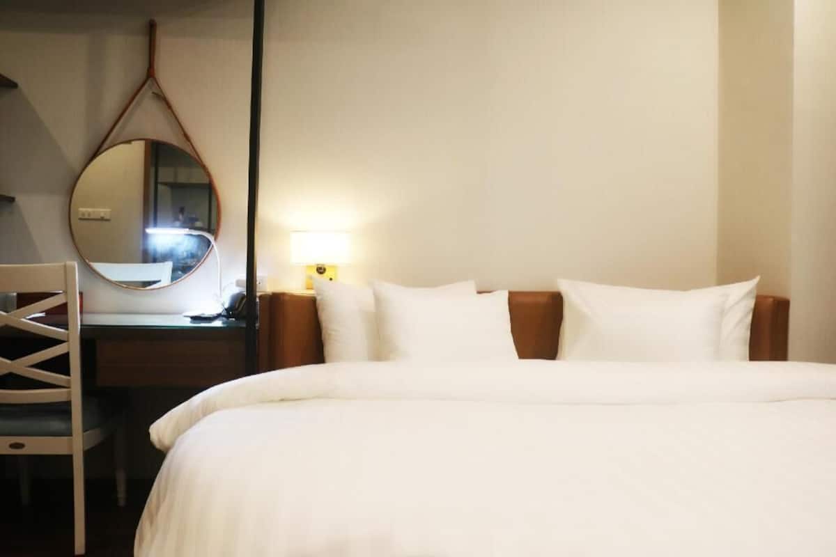 A hotel room with a bed, a desk with a chair, a round mirror, and a bedside lamp.