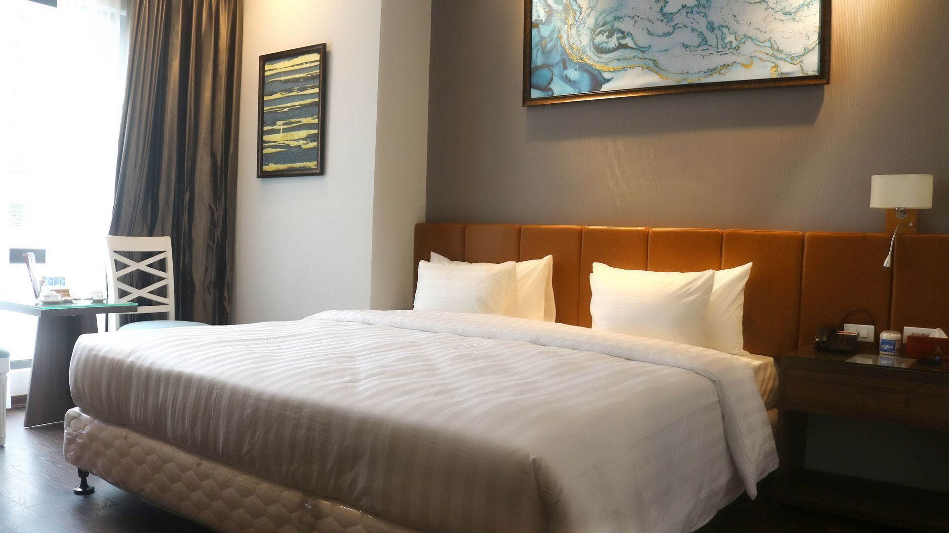 A hotel room with a large bed, a headboard, a nightstand, a lamp, a painting on the wall, and a window with curtains.