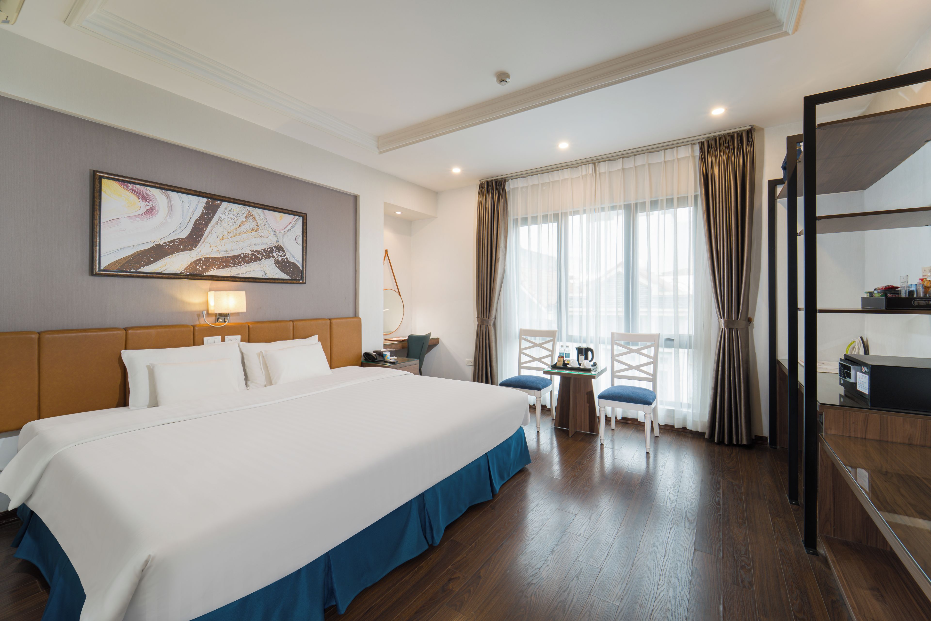 Ein Hotelzimmer mit einem großen Bett, einem Schreibtisch, einem Stuhl, einem fenster mit Vorhängen und einem Bild an der Wand.