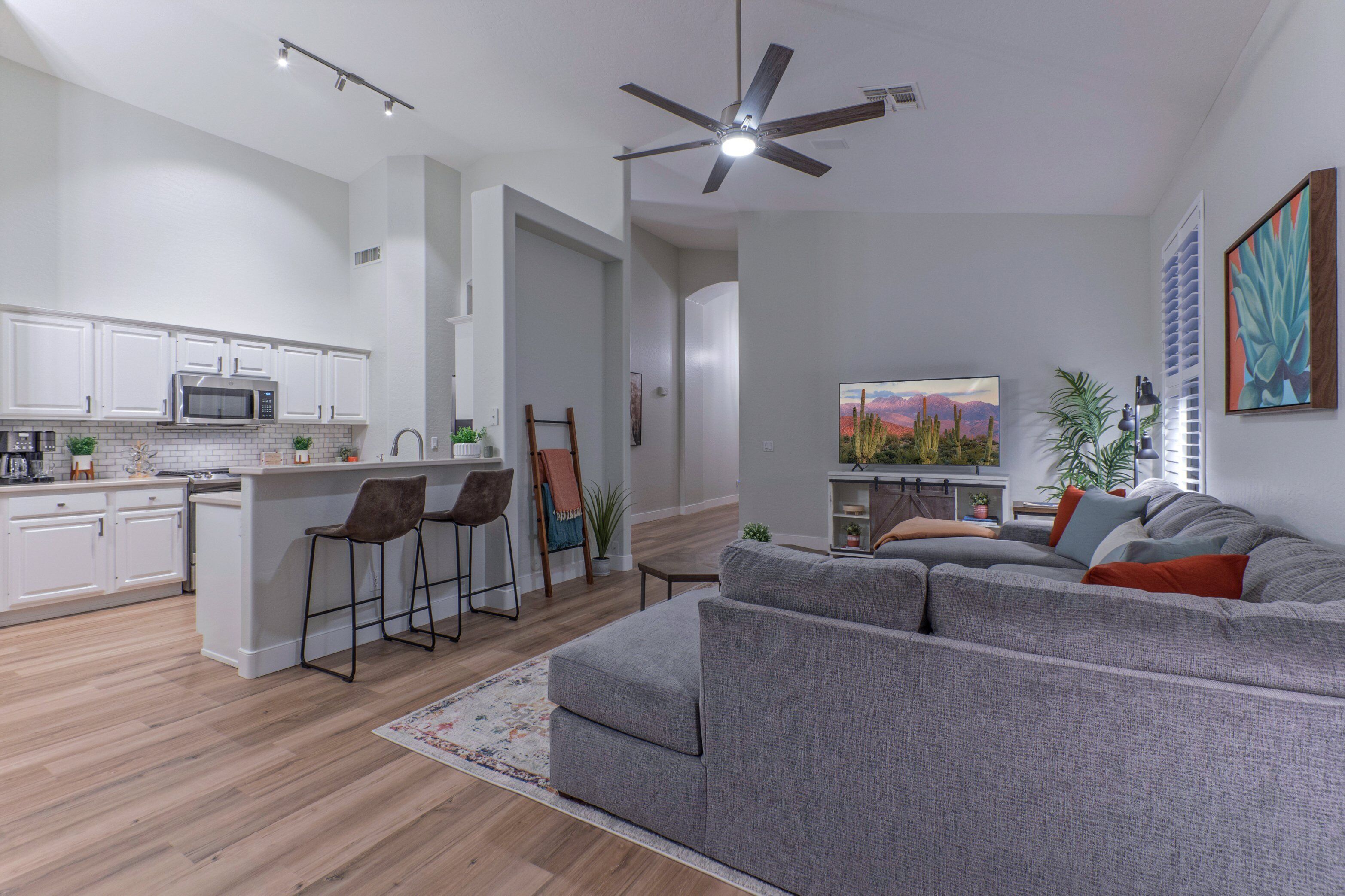 A modern kitchen with white cabinets, a microwave, and a bar stool. There is a gray sofa with cushions in the living area.