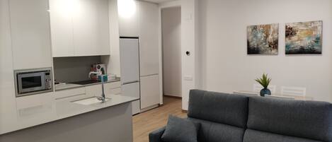 A modern kitchen with white cabinets, a built-in microwave, and a sink. There is a grey sofa in the living area with a small table and a plant. Two abstract paintings are on the wall.