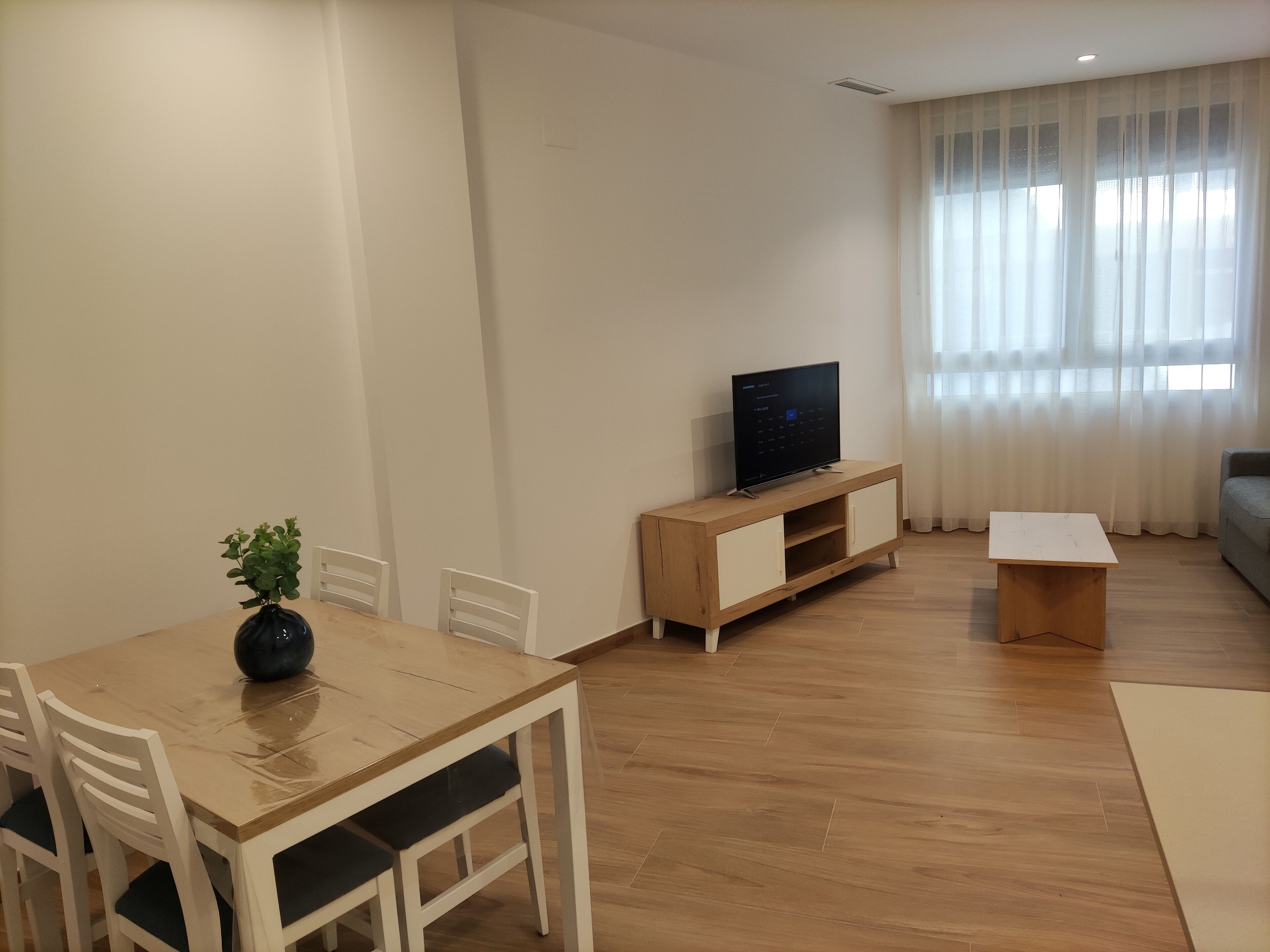 Una sala de estar moderna con una mesa de comedor de madera, sillas, un televisor sobre un mueble de madera, una mesita blanca pequeña y un ventanal con cortinas de encaje.