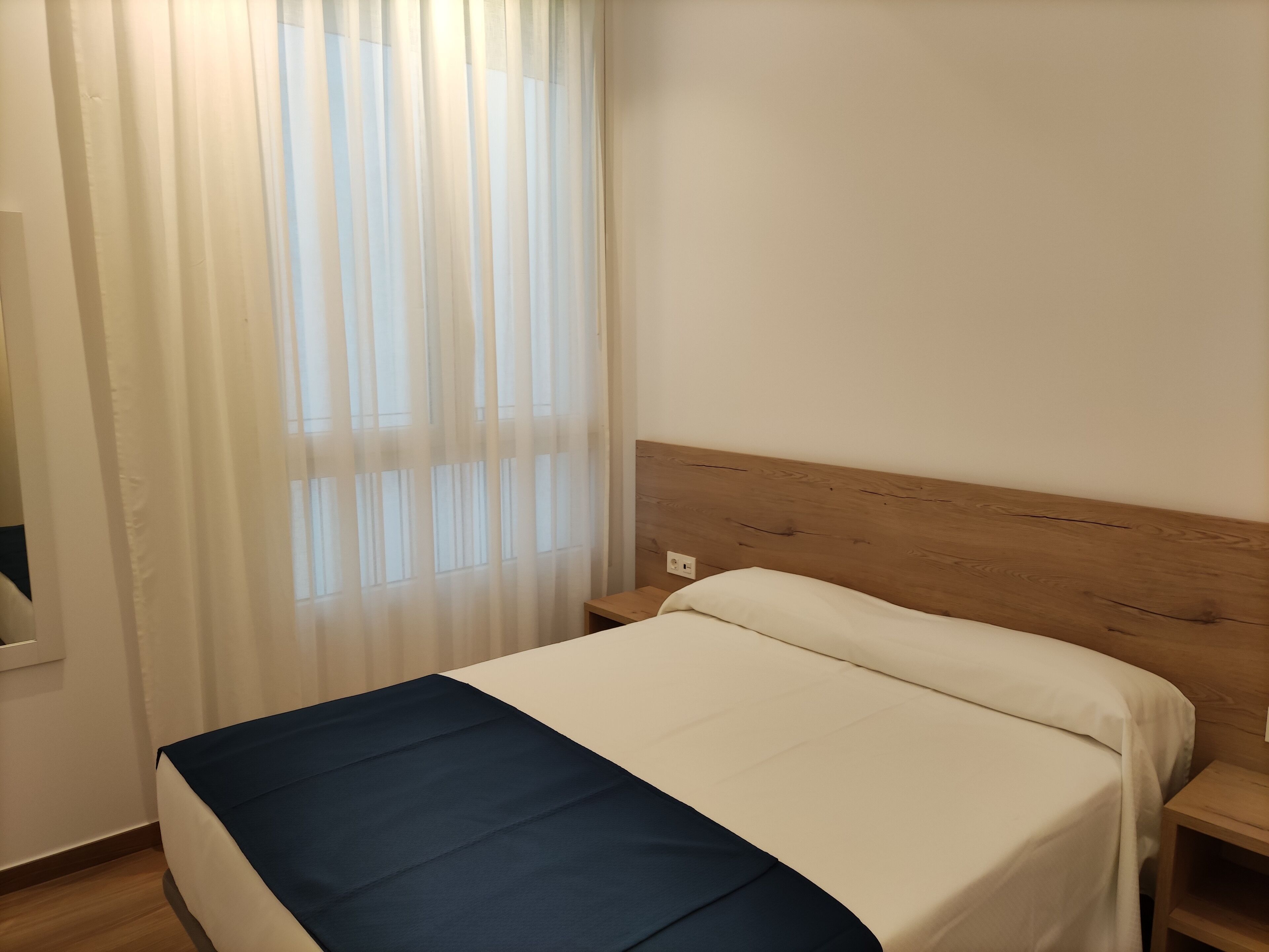Una cama individual con cabecera de madera, colcha blanca y azul, y una ventana con cortinas traslúcidas.