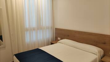 A single bed with a wooden headboard, a white and blue bedspread, and a window with sheer curtains.
