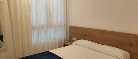 A single bed with a wooden headboard, a white and blue bedspread, and a window with sheer curtains.