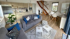 Ein modernes Wohnzimmer mit grauem Sofa, Couchtisch und Essbereich mit weißem Tisch und Stühlen. Im Hintergrund ist eine Treppe zu sehen.
