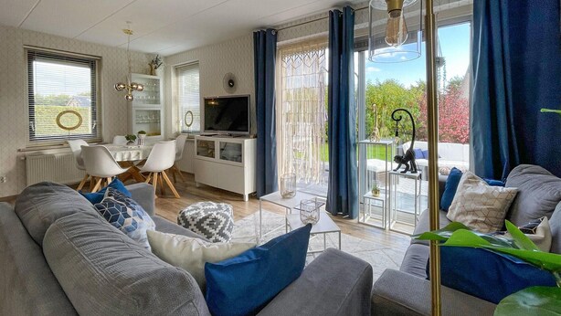 Ein modernes Wohnzimmer mit Sofa, Couchtisch und Fernseher. Es gibt einen Essbereich mit Stühlen und Tisch. Große Fenster mit Vorhängen bieten einen Blick ins Freie.