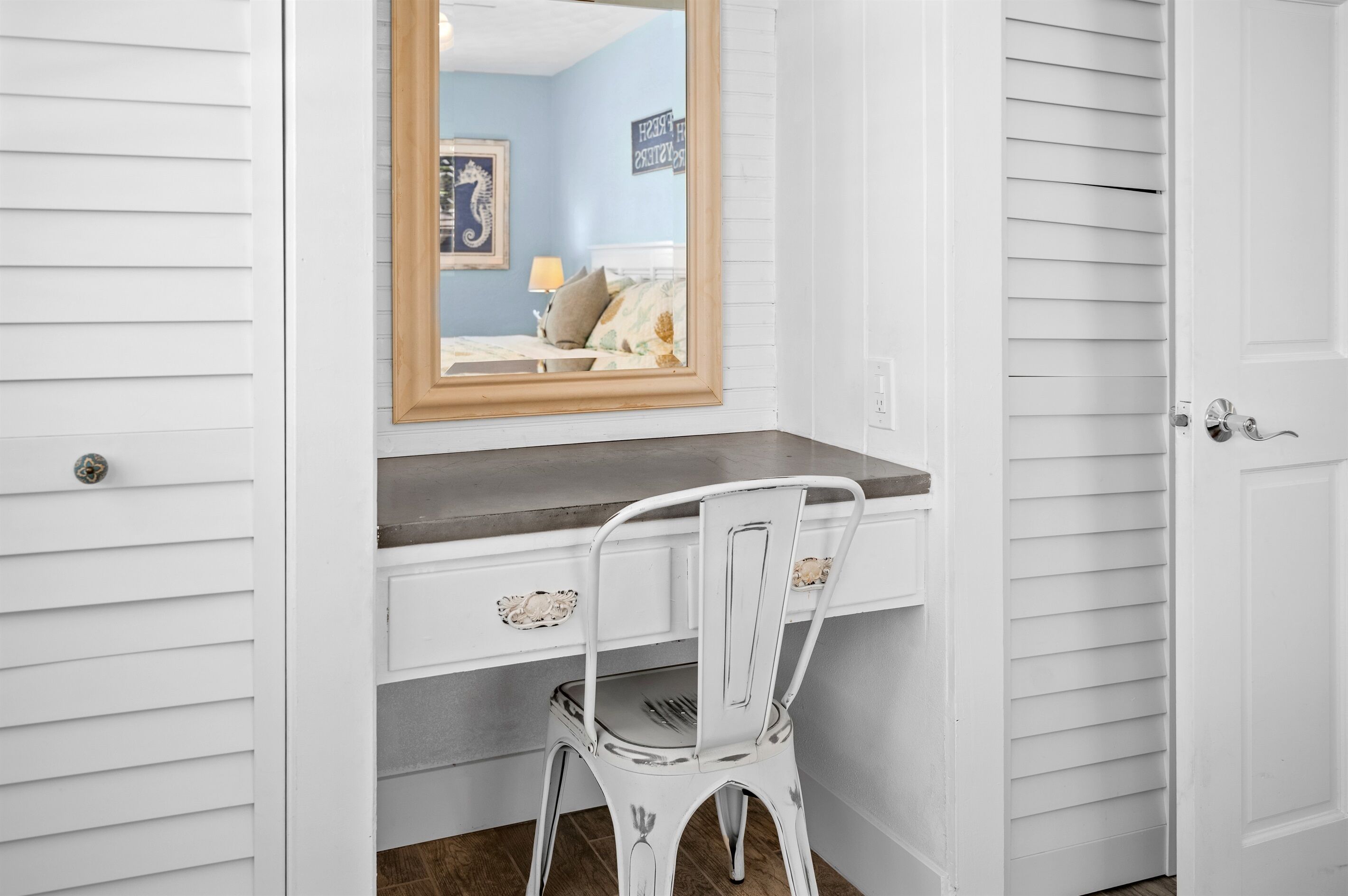 Une pièce blanche avec un miroir à cadre en bois au-dessus d’un bureau blanc, une chaise blanche et une porte avec une poignée argentée.