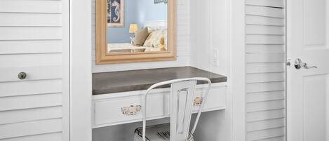 Une pièce blanche avec un miroir à cadre en bois au-dessus d’un bureau blanc, une chaise blanche et une porte avec une poignée argentée.