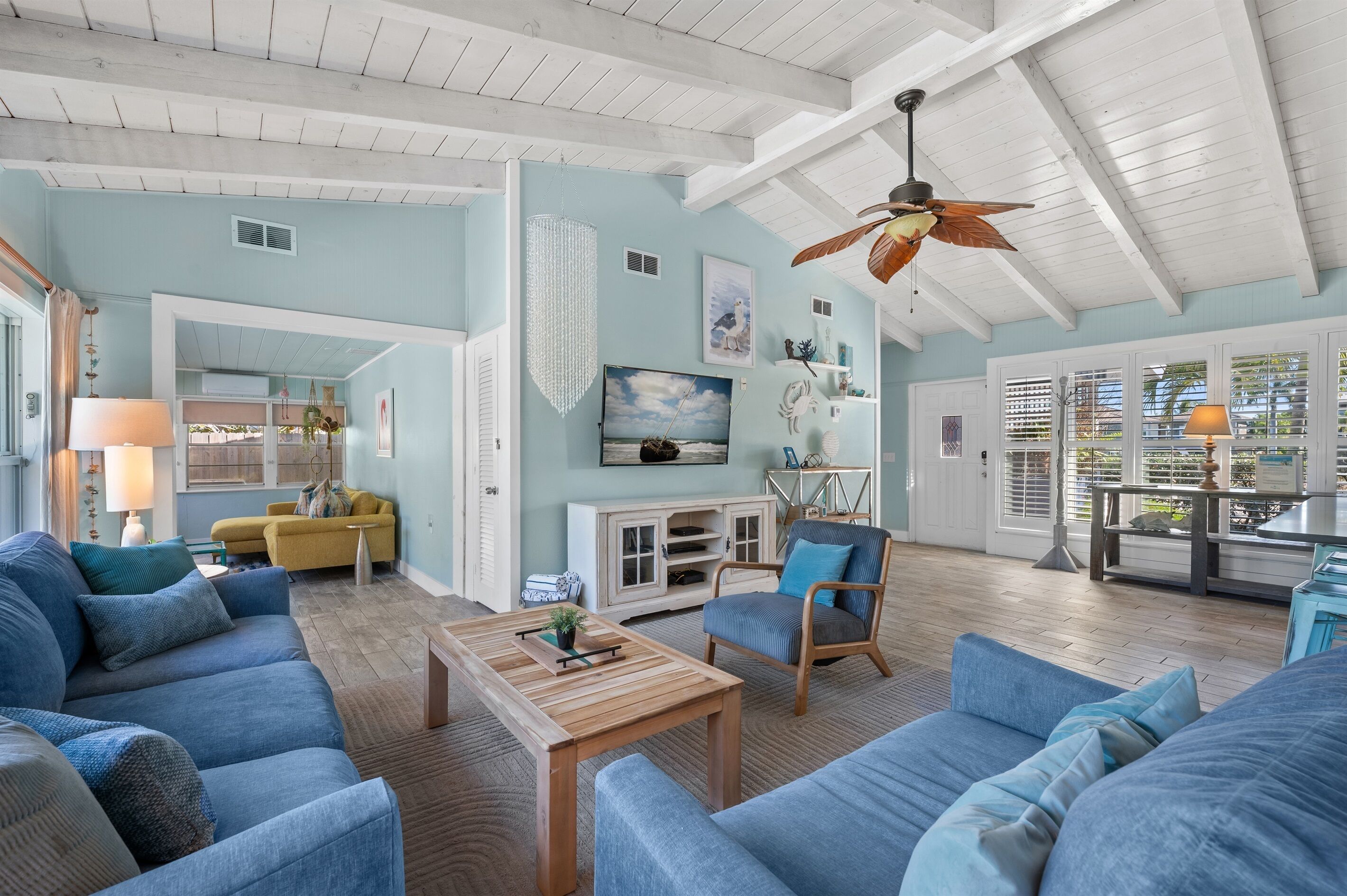 A spacious living room with a high ceiling, a wooden ceiling fan, and a large window with blinds. There is a blue sofa, a wooden coffee table, and a chair with blue cushions.